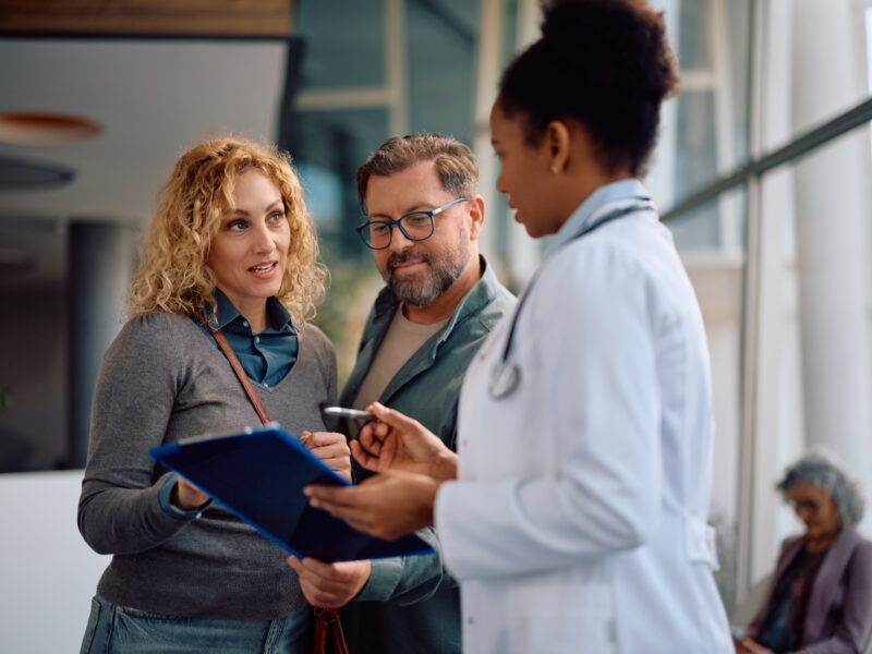 Mid,Adult,Woman,With,Husband,Talking,To,A,Doctor,About